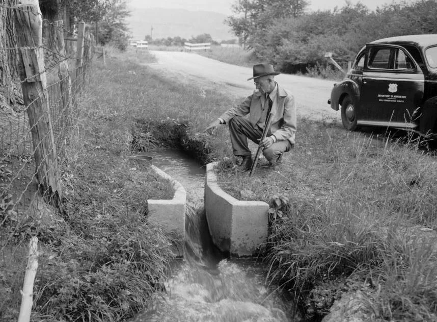Ralph Parshall measuring water in Parshall flume with Soil Conservation Service vehicle in background.