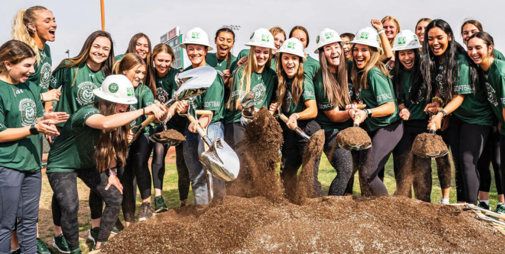 Women's soccer and softball teams wearing helmets and holding shovels for the ground breaking ceremony of the new women's sports complex
